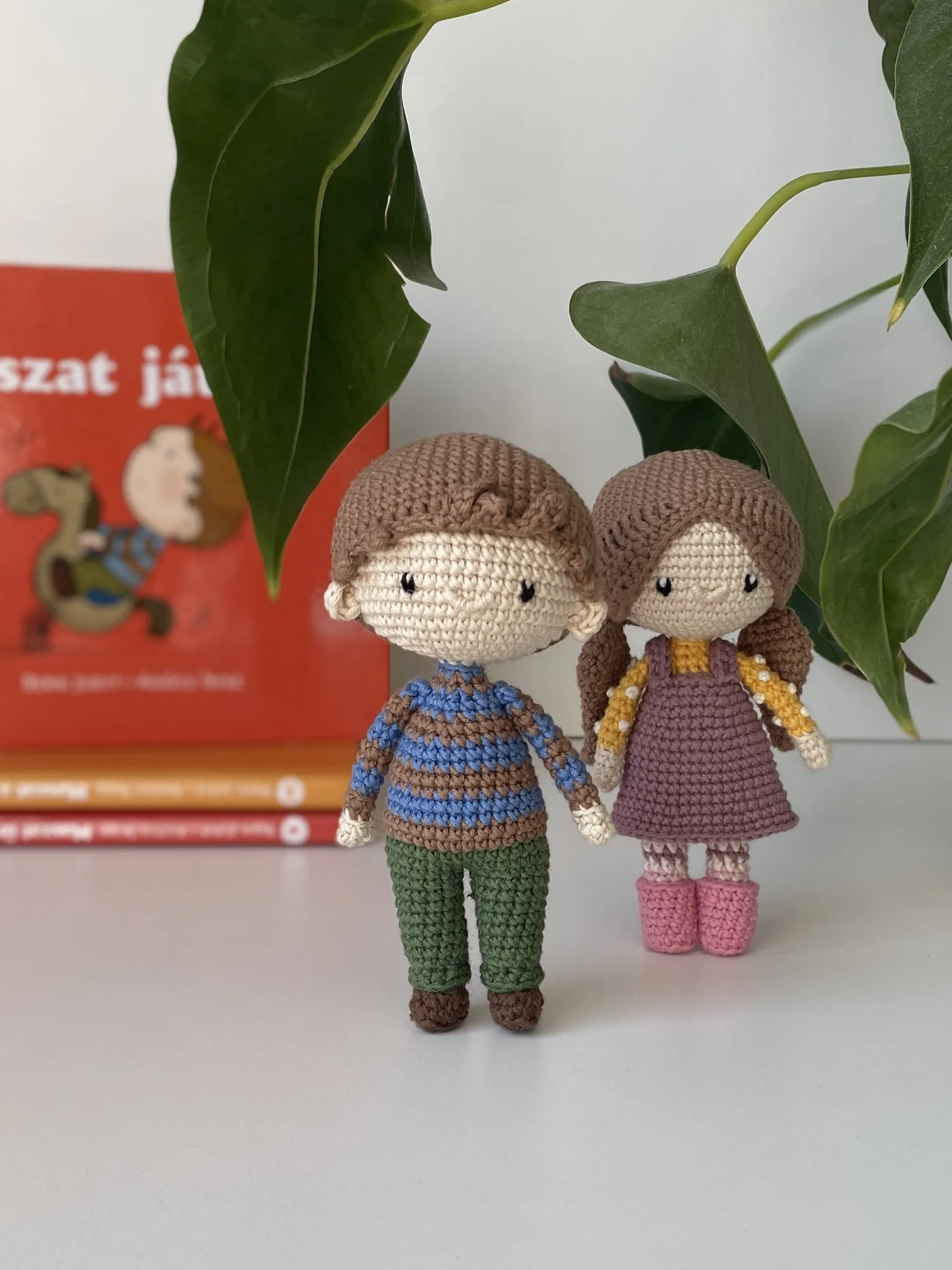 Kézzel horgolt Maszat és Sári figura egymás mellett, mögöttük egy Maszat-könyv és zöld levelek